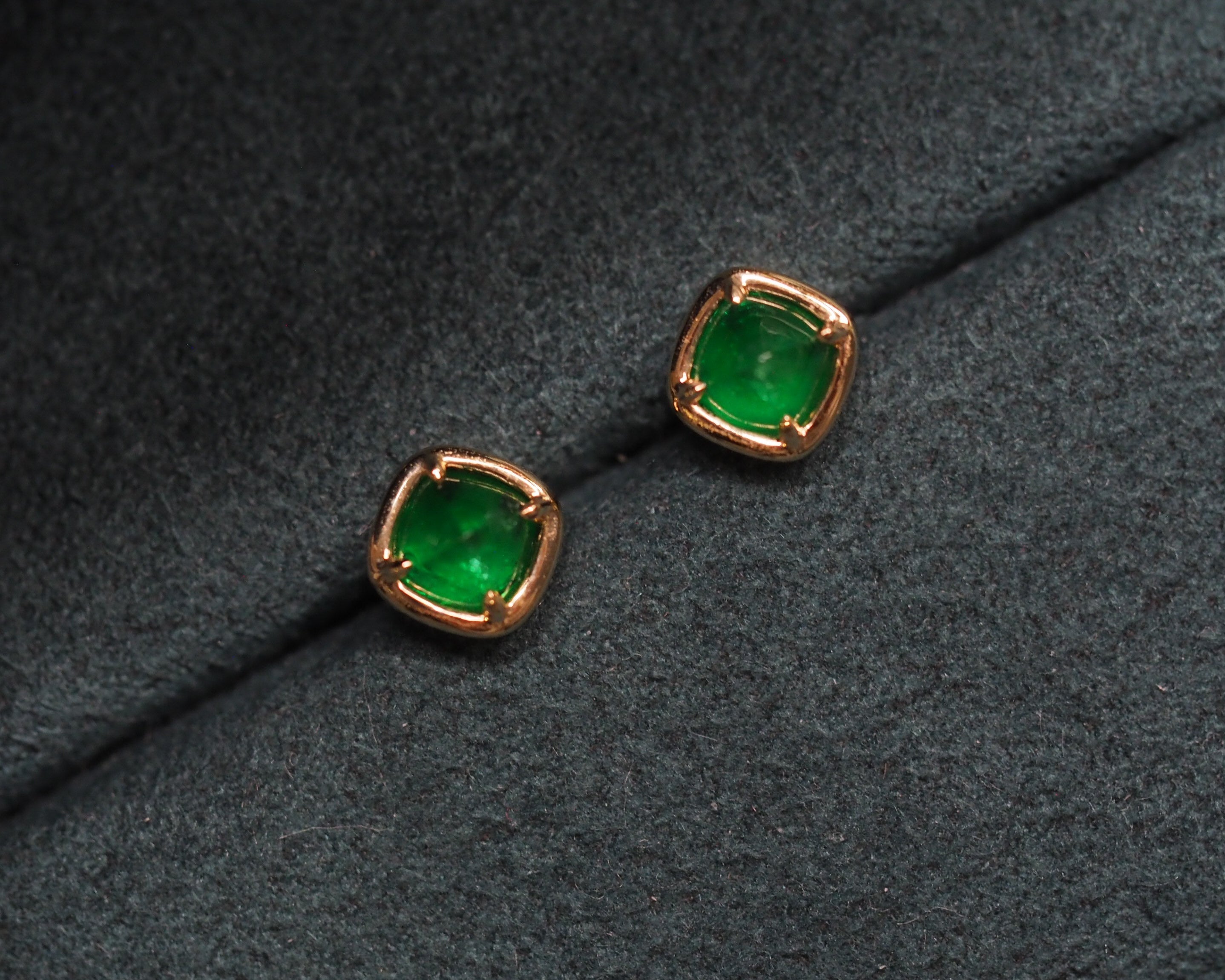 A pair of sugarloaf-cut green emerald stud earrings with gold settings, displayed on a textured dark gray velvet surface.