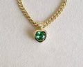A gold chain necklace with a heart-shaped pendant featuring a vibrant green heart-shaped gemstone, displayed on a textured off-white background.