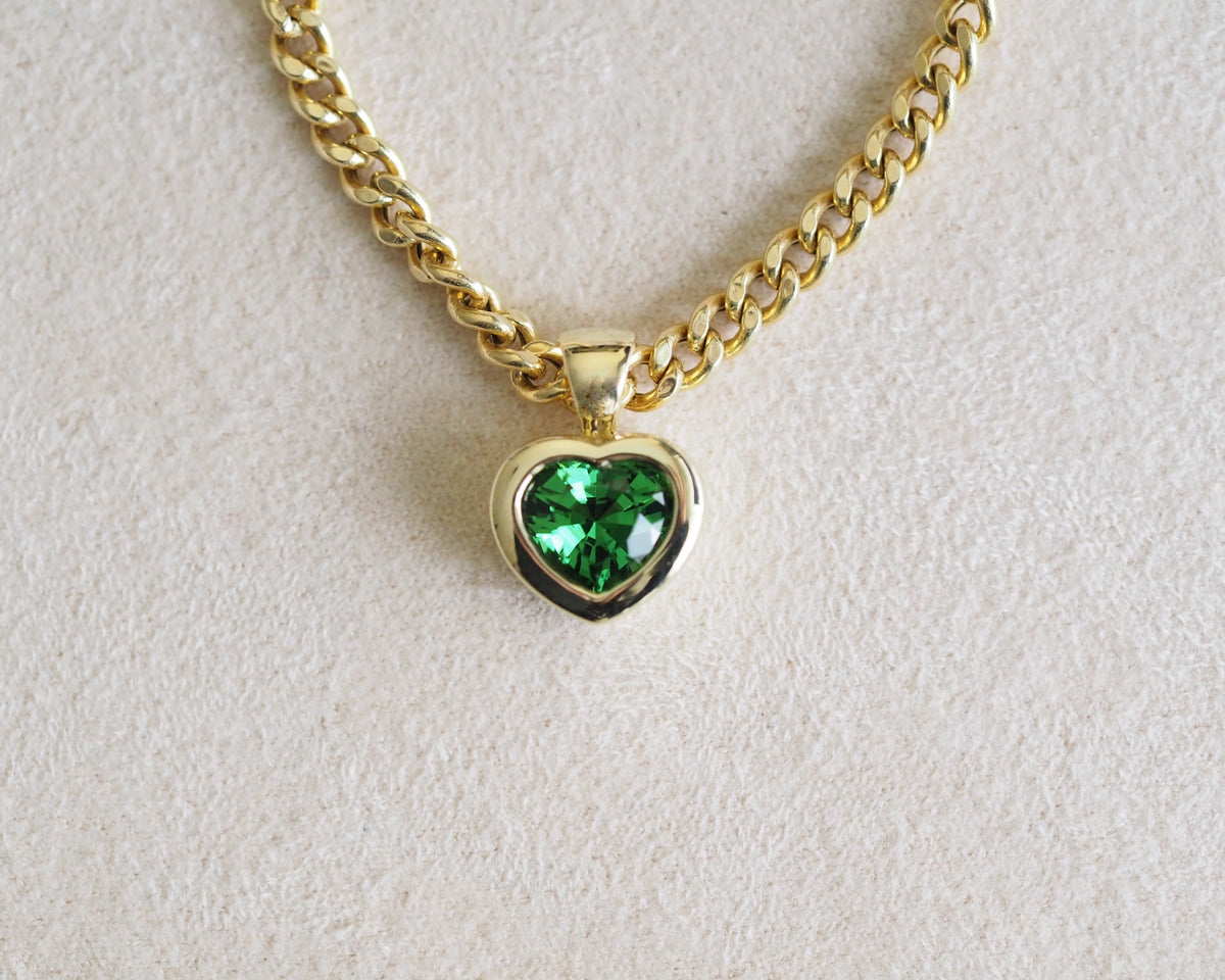 A gold chain necklace with a heart-shaped pendant featuring a vibrant green heart-shaped gemstone, displayed on a textured off-white background.