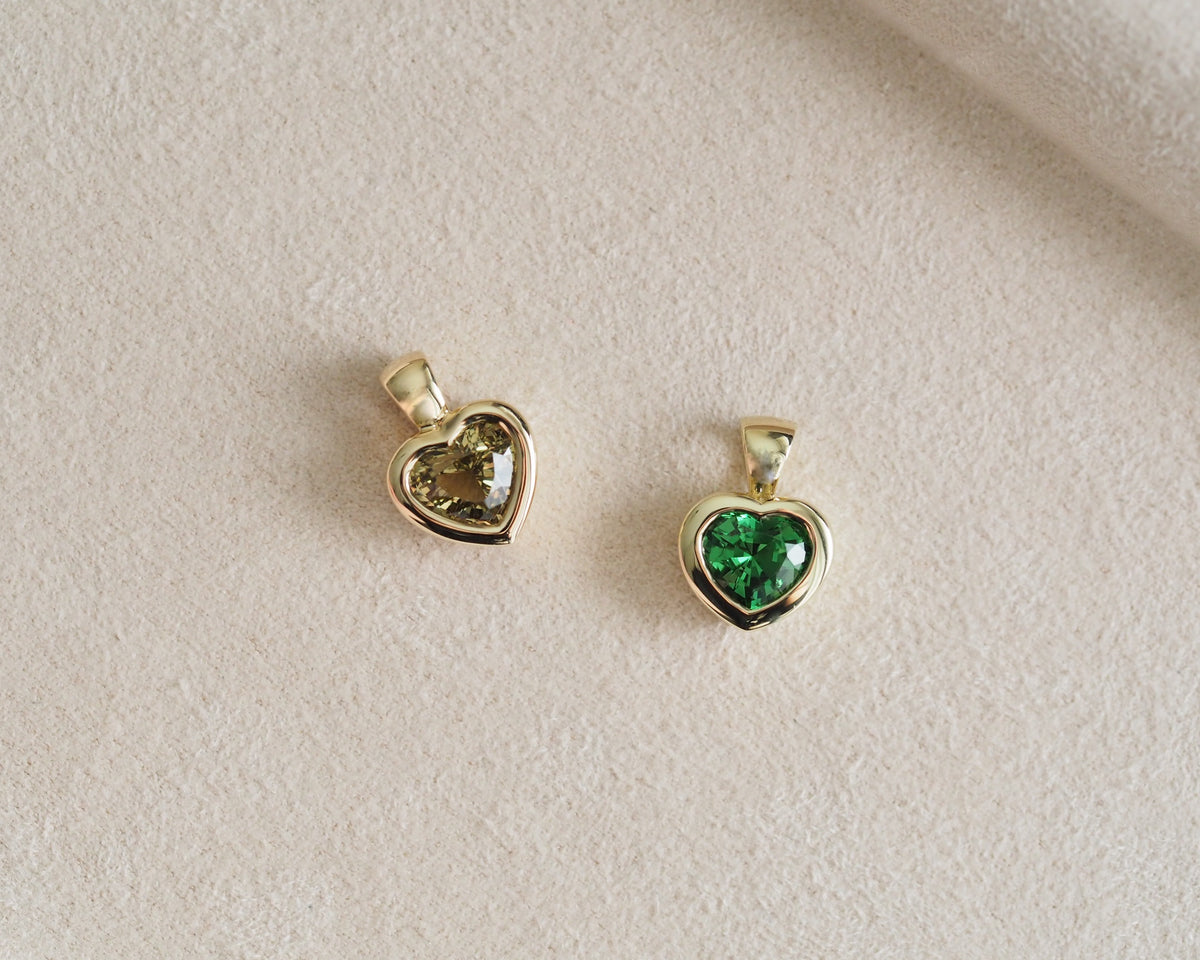 Two gold heart-shaped pendant charms on a textured beige surface, one with a yellow-green heart-shaped gemstone and the other with a vivid green heart-shaped gemstone.