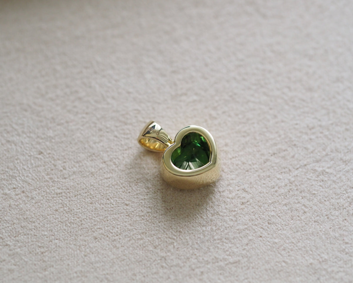 A gold heart-shaped pendant with a faceted green gemstone, resting on a textured beige fabric background.