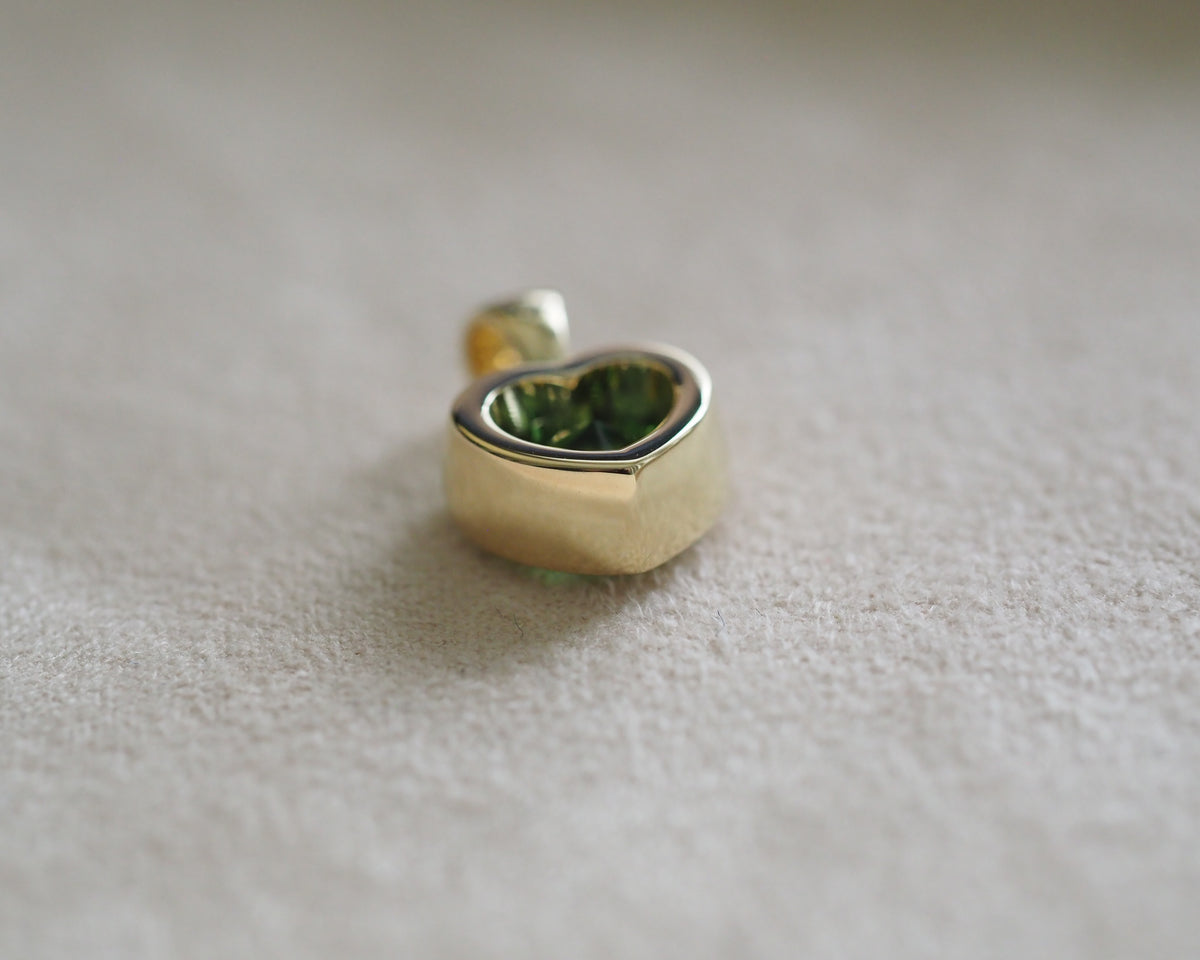 A small gold heart-shaped stud earring with a polished finish and a green recessed interior, resting on a soft beige textured surface.