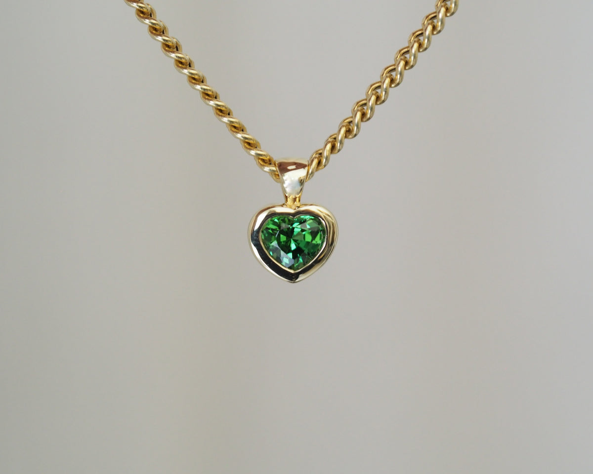 A gold heart-shaped pendant necklace featuring a faceted green gemstone, suspended from a twisted gold chain against a neutral light gray background.