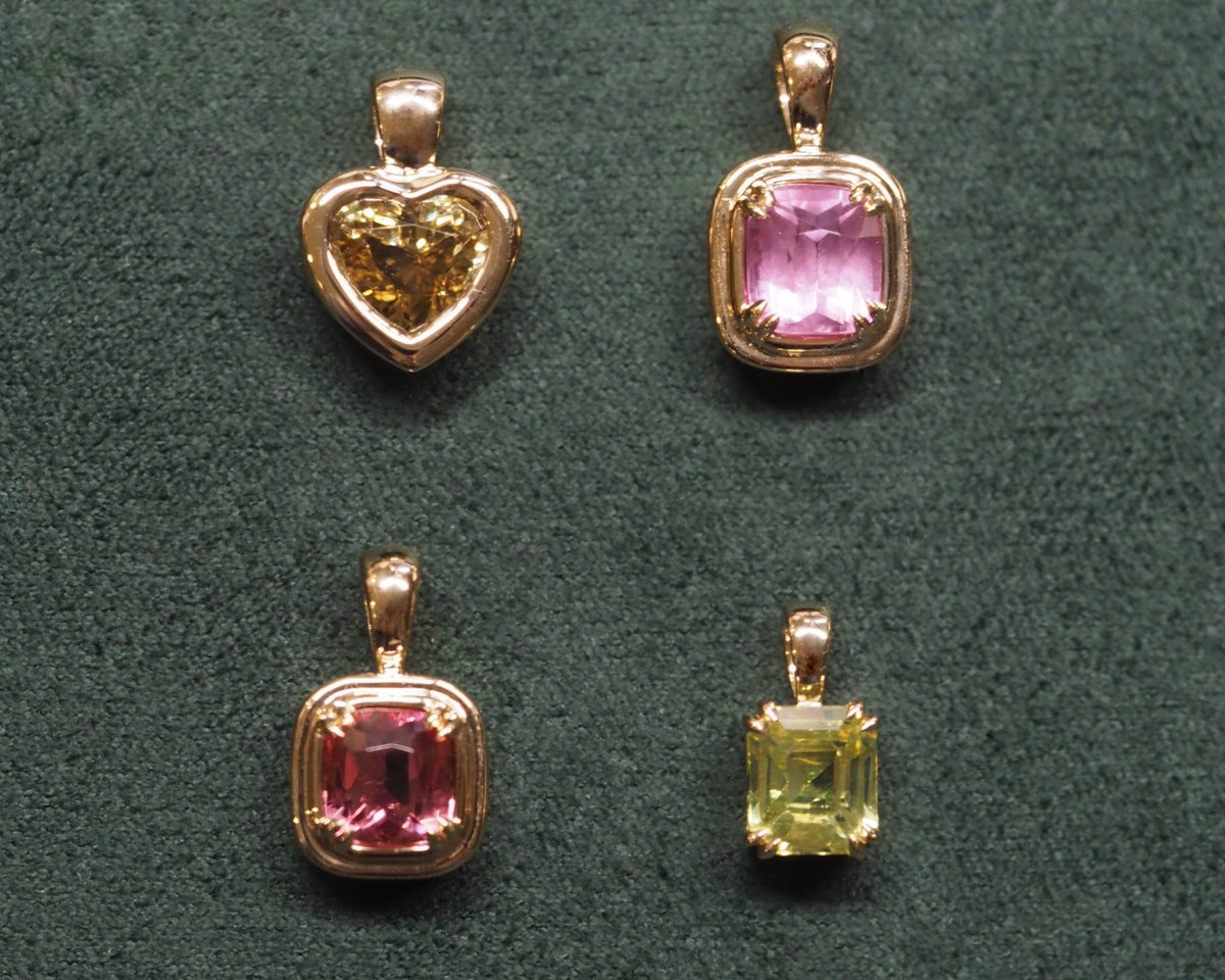 Four gold-toned gemstone pendants arranged on a dark green textured background, including a heart-shaped pendant with a yellow gemstone and three square pendants with pink and light green gemstones.