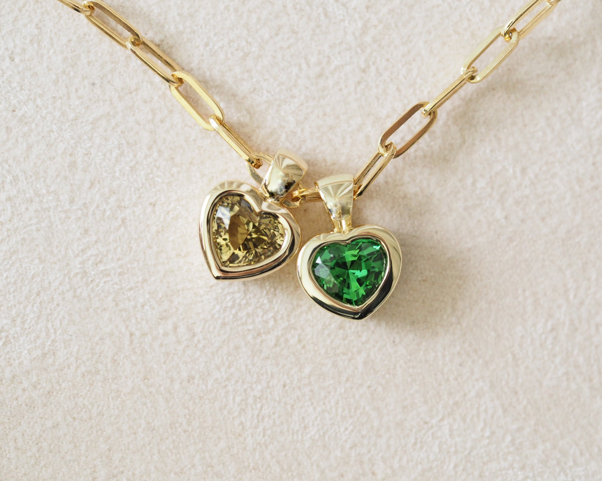 A gold paperclip chain necklace with two heart-shaped pendants, one featuring a yellow gemstone and the other a green gemstone, displayed on a textured off-white background.