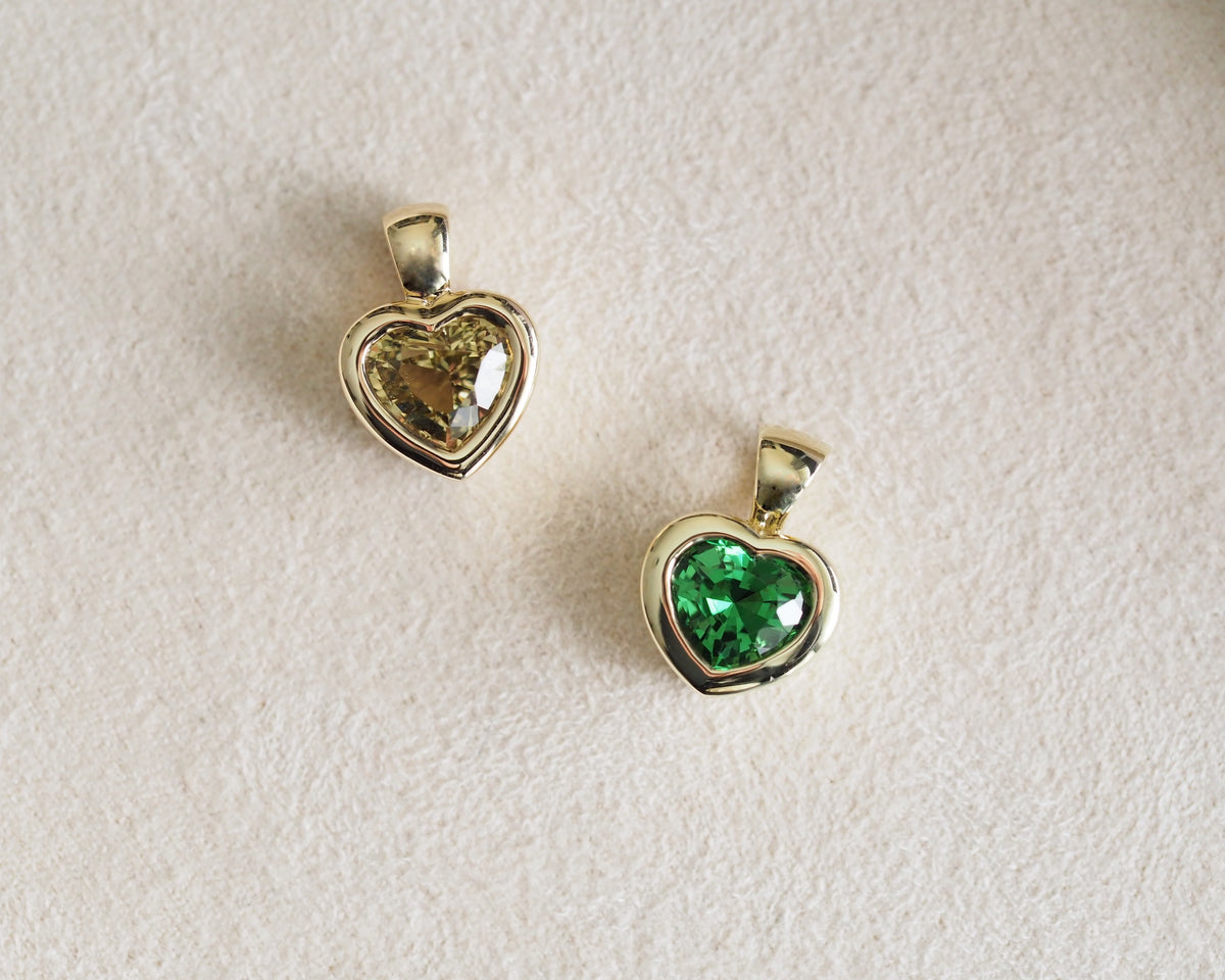 Two gold heart-shaped pendant charms on a textured off-white background. One pendant features a clear faceted gemstone, while the other features a vibrant green faceted gemstone, both set in polished gold-tone metal.