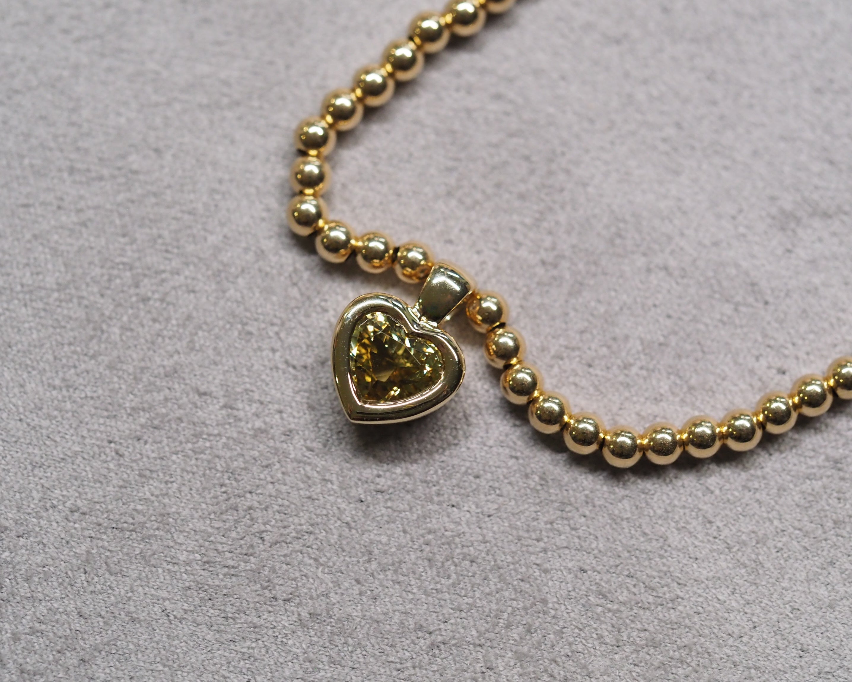 A gold beaded necklace with a heart-shaped pendant containing a yellow-green heart-shaped stone, displayed on a textured gray background.