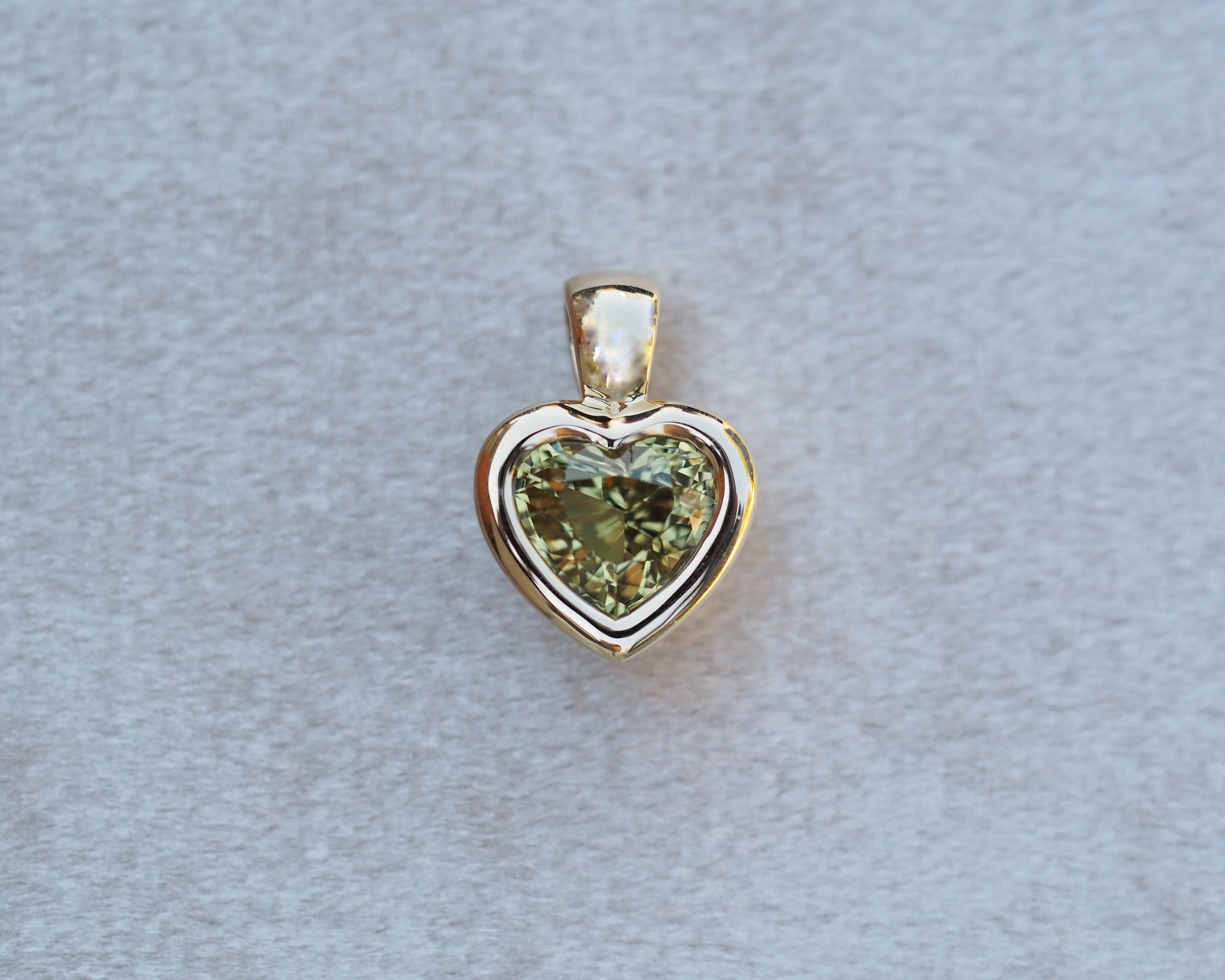 A heart-shaped pendant featuring a faceted olive green gemstone in a polished yellow gold setting, displayed on a textured light gray background.