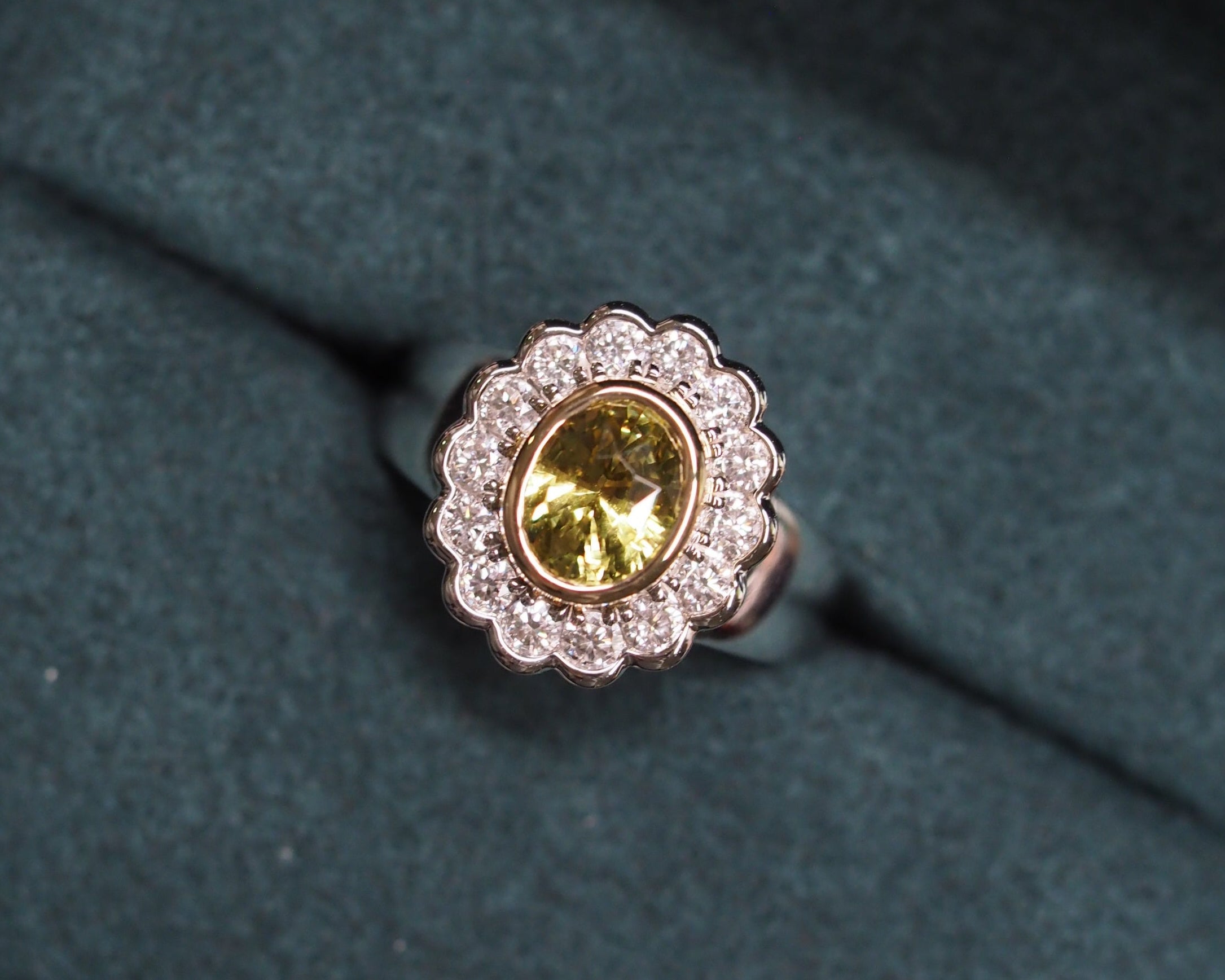 A two tone gold ring with a scalloped halo of small clear stones and a central oval yellow chrysoberyl, displayed on a dark textured velvet surface.