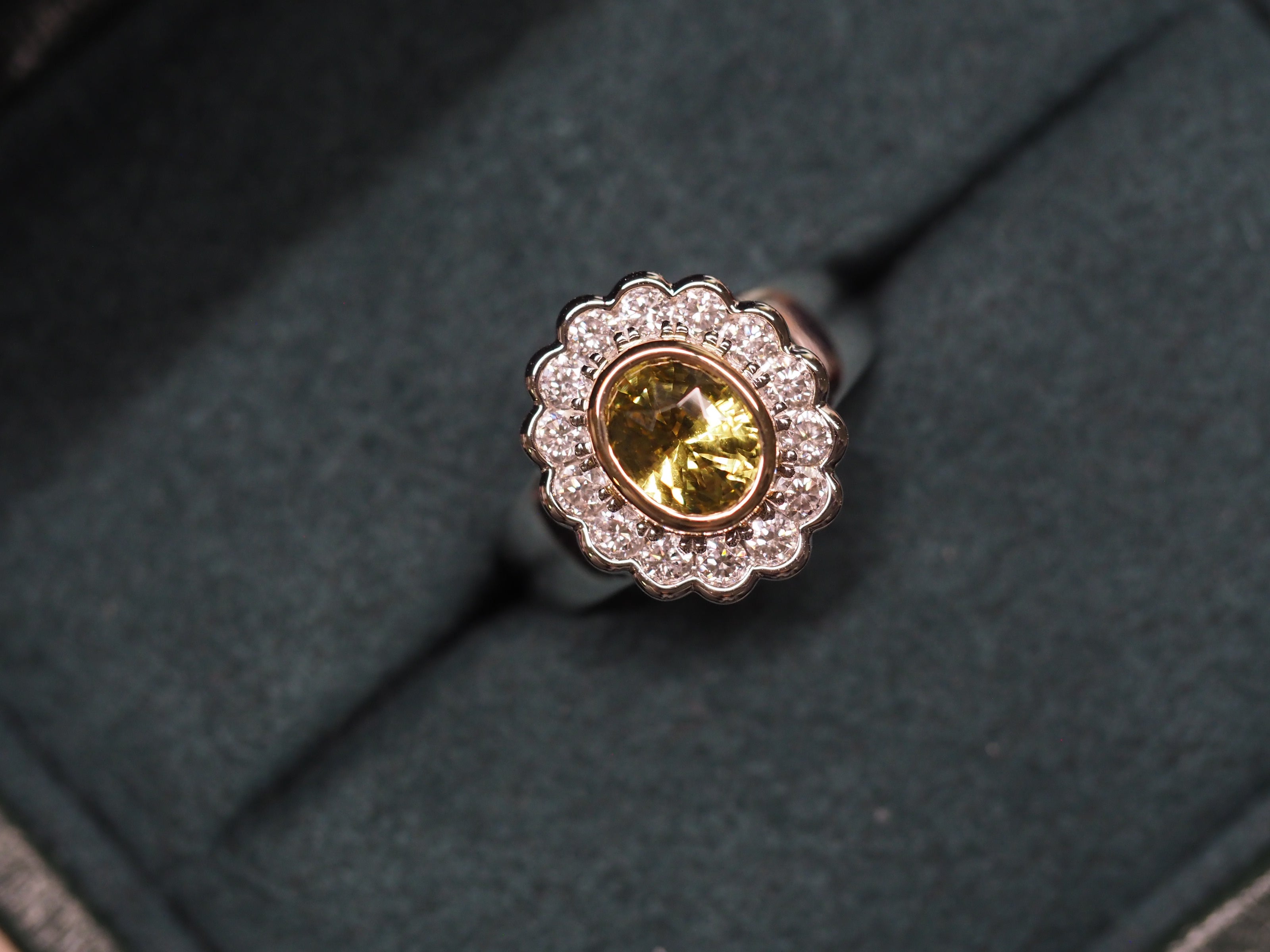 A two tone gold ring with a yellow oval chrysoberyl and a halo of small clear stones, displayed on a dark textured velvet ring box.