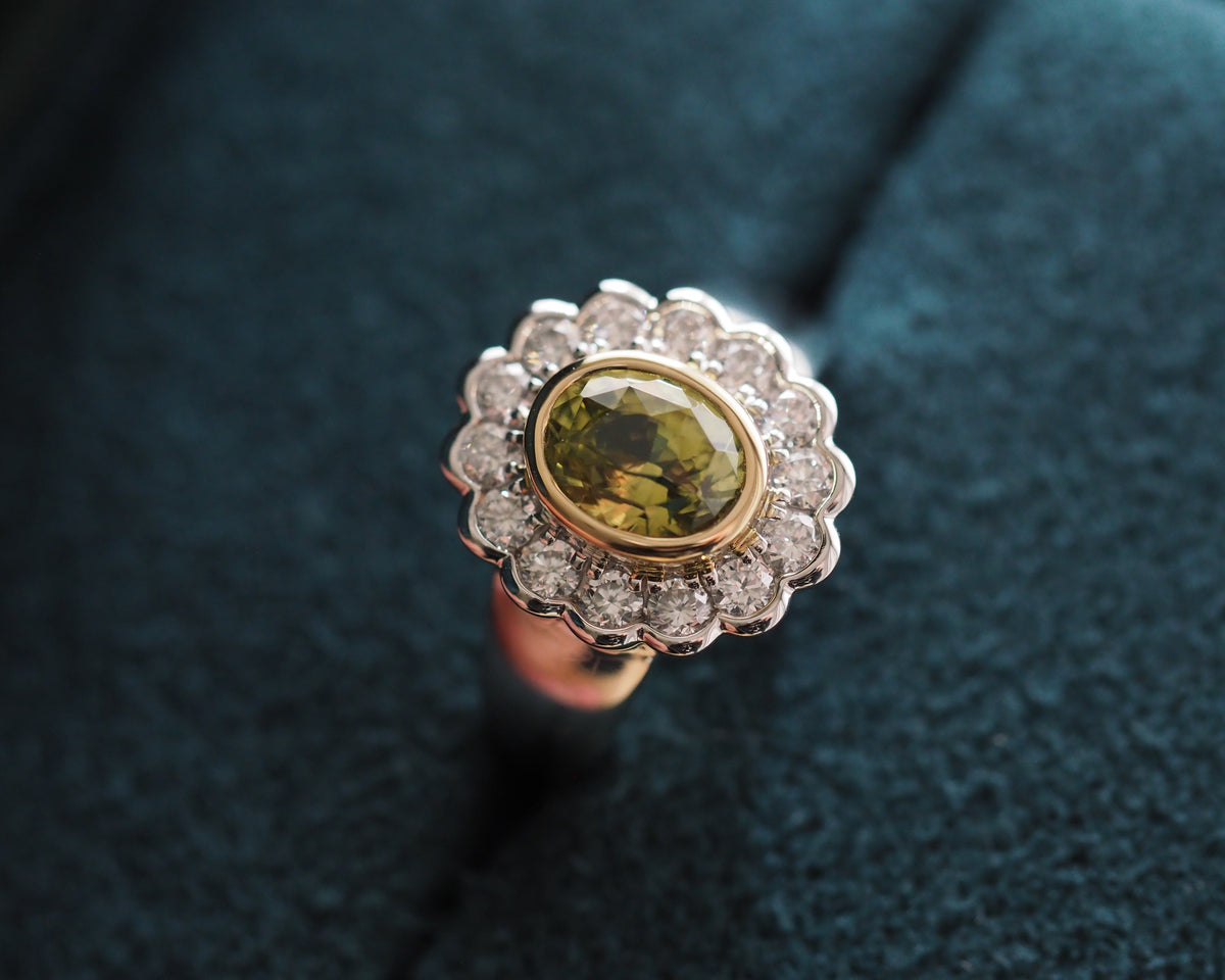 A close-up of a vintage-style ring featuring a central oval olive-green gemstone surrounded by a halo of small clear stones, set in a yellow-gold band, resting on a dark teal velvet surface.