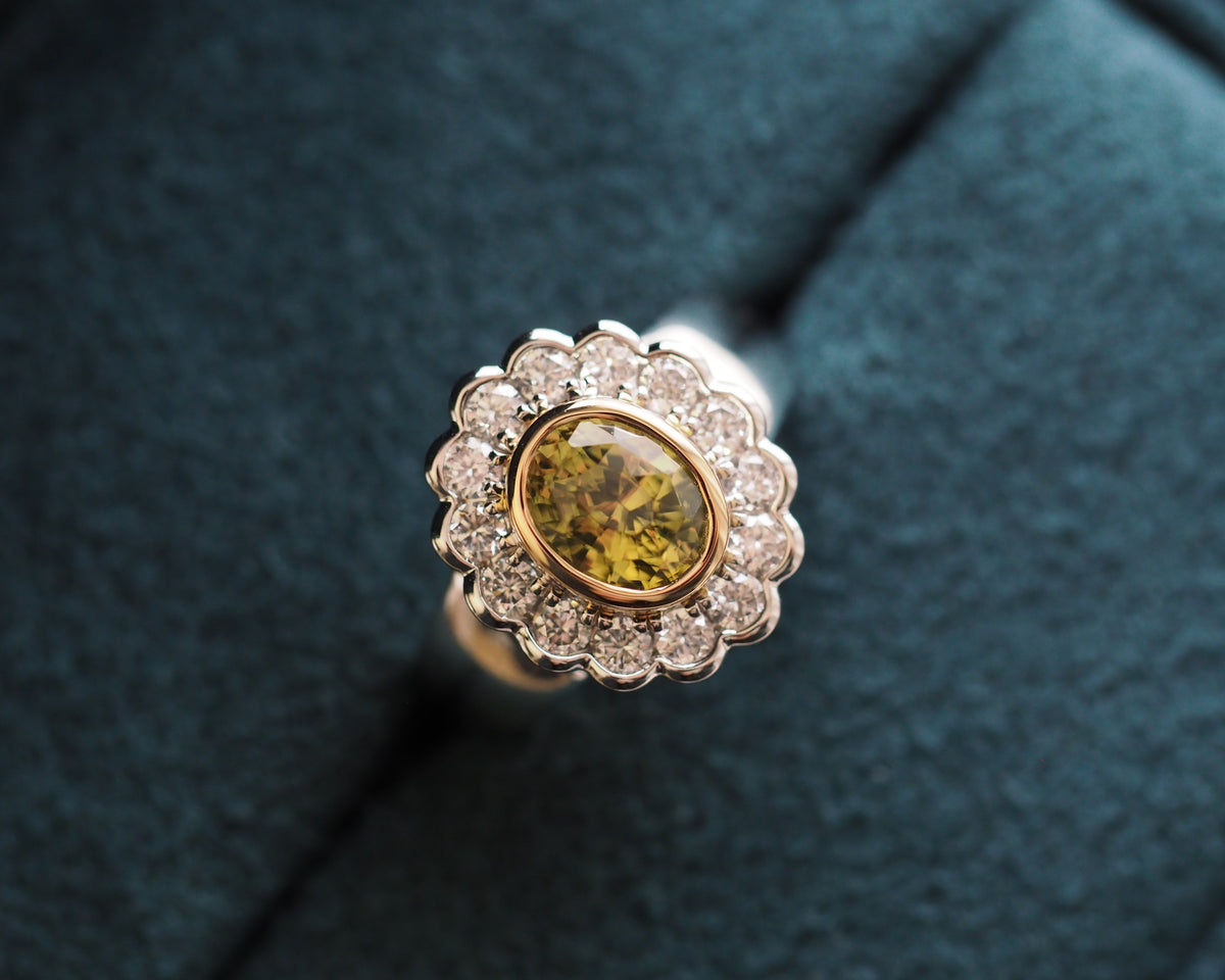 A close-up of a vintage-style ring featuring a yellow diamond center stone surrounded by a halo of small clear stones in a gold-toned setting, displayed on a dark teal velvet ring box.