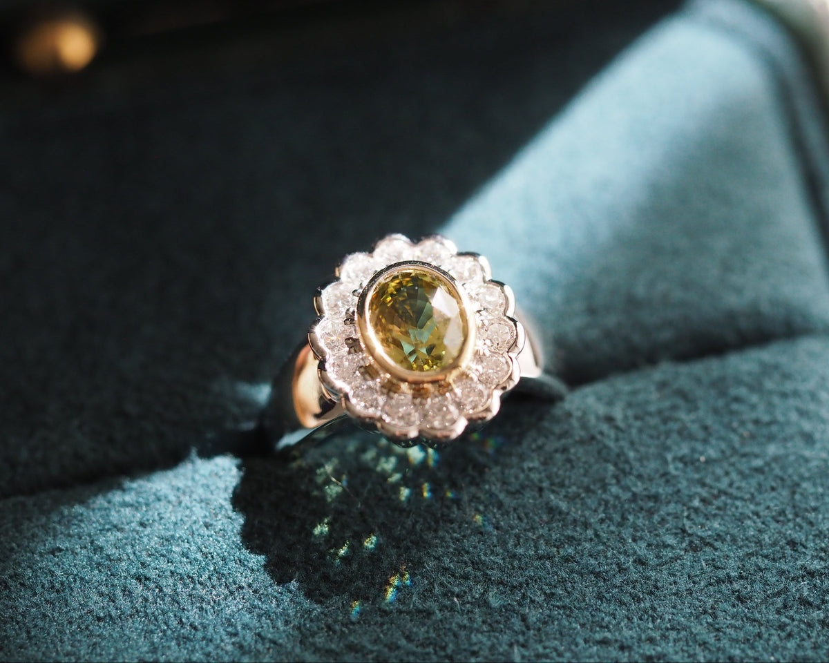 A vintage-style ring with a scalloped silver-toned halo and a central round yellow-green stone, displayed on a dark teal velvet ring box.
