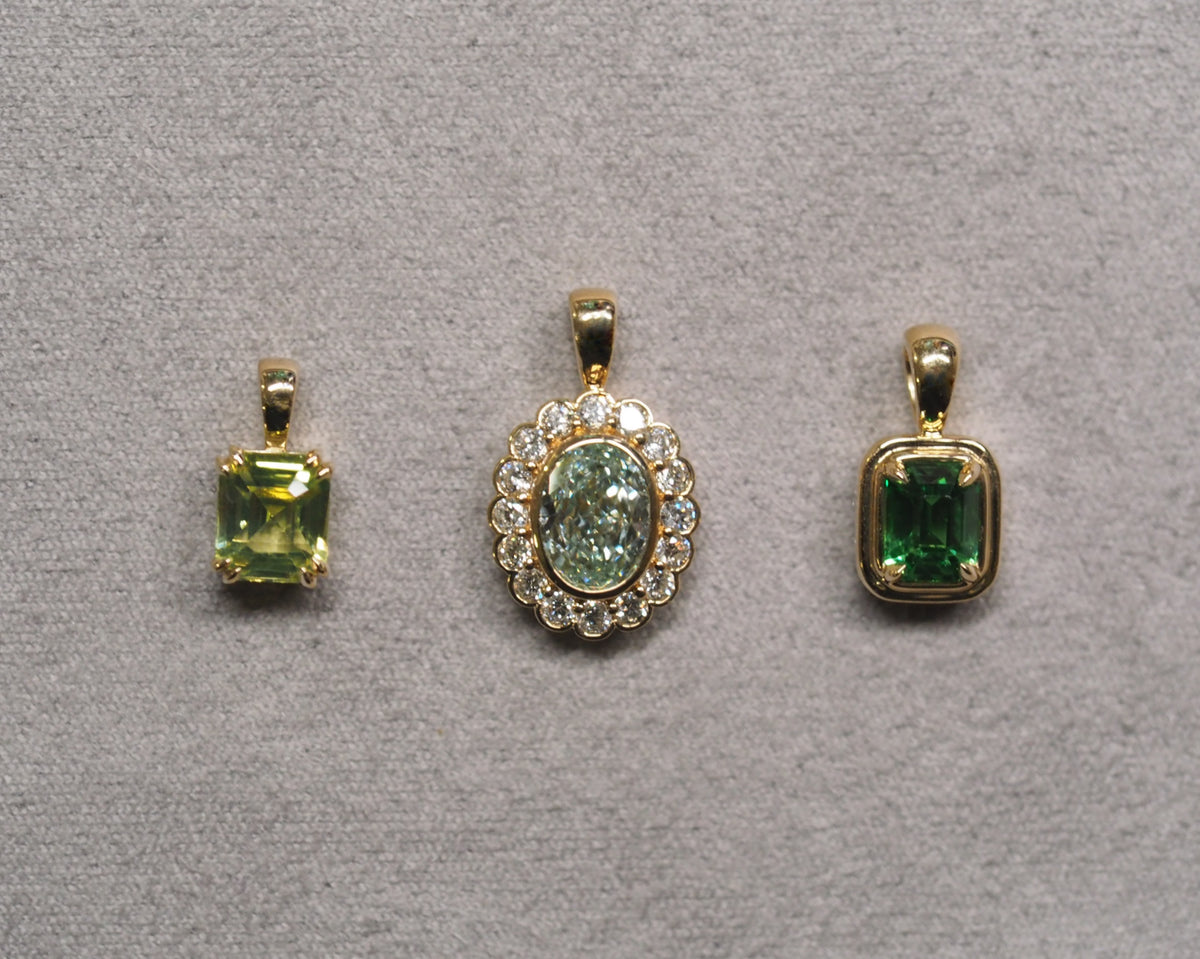 Three gold pendant necklaces displayed on a textured gray fabric background. The left pendant features a rectangular green gemstone in a gold bezel setting.