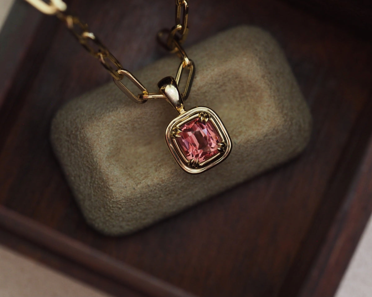 A gold chain necklace with a square bezel-set pink Malaya garnet pendant, displayed on a textured olive-green jewelry cushion over a dark wooden surface.