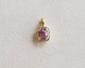 A gold pendant featuring a square-cut, cushion-shaped pinkish-purple garnet, displayed on a textured off-white background.