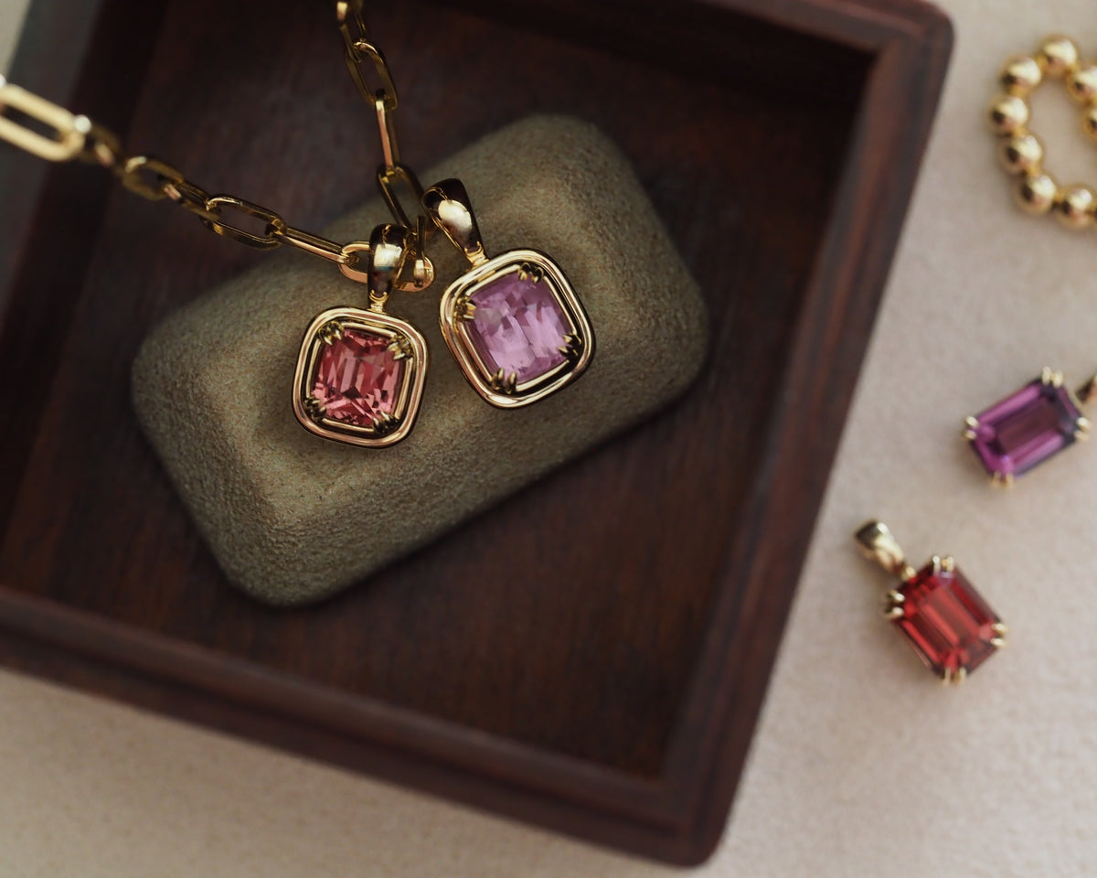 Gold-toned jewelry set featuring a necklace and earrings with rectangular pink gemstones, displayed on a dark wooden tray with a gray cushion. Additional matching jewelry pieces are arranged on a light beige surface nearby.