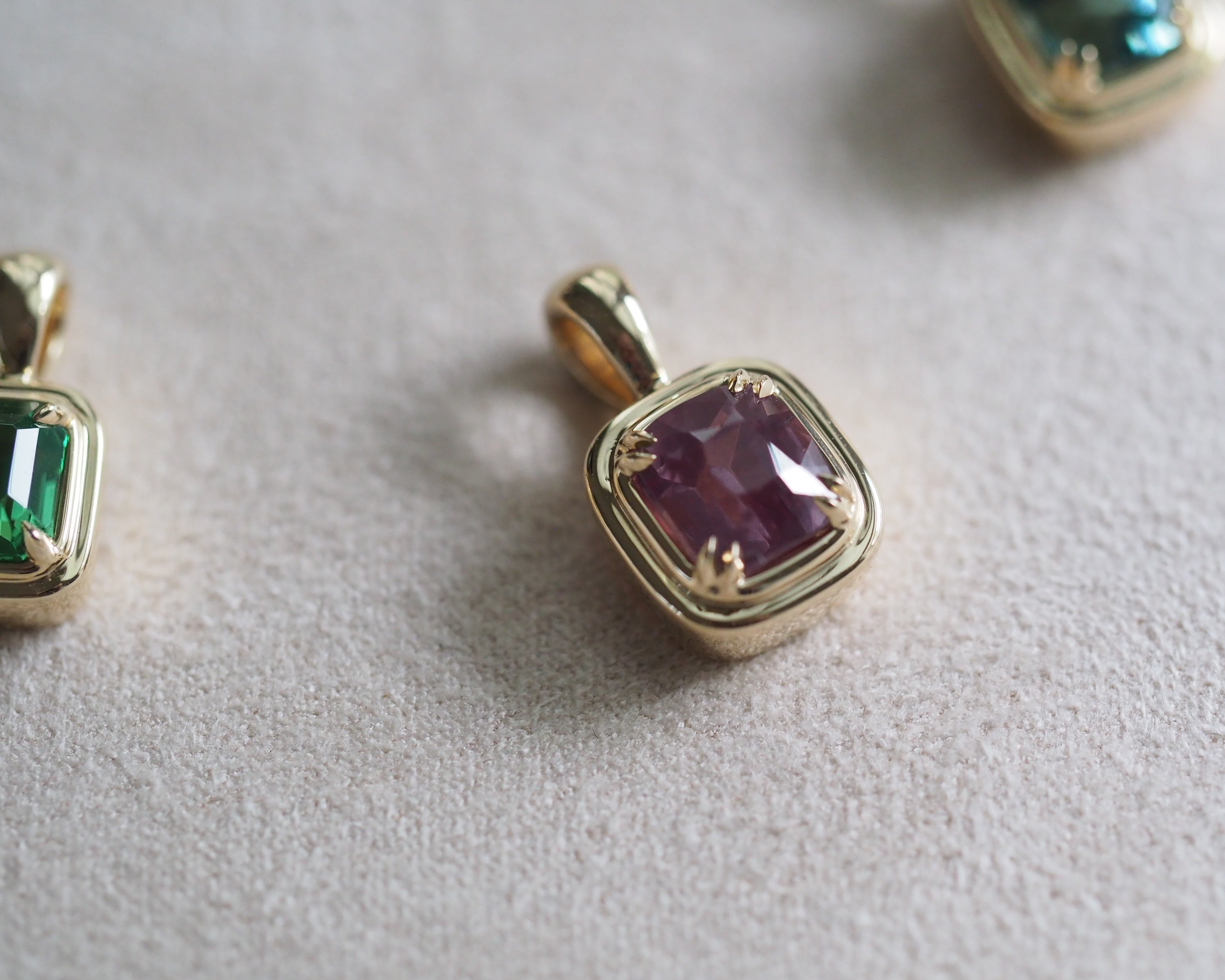 A gold pendant with a cushion-cut milky garnet, shown in close-up on a textured light beige surface with Robyn and Juno pendant visible at the edges.
