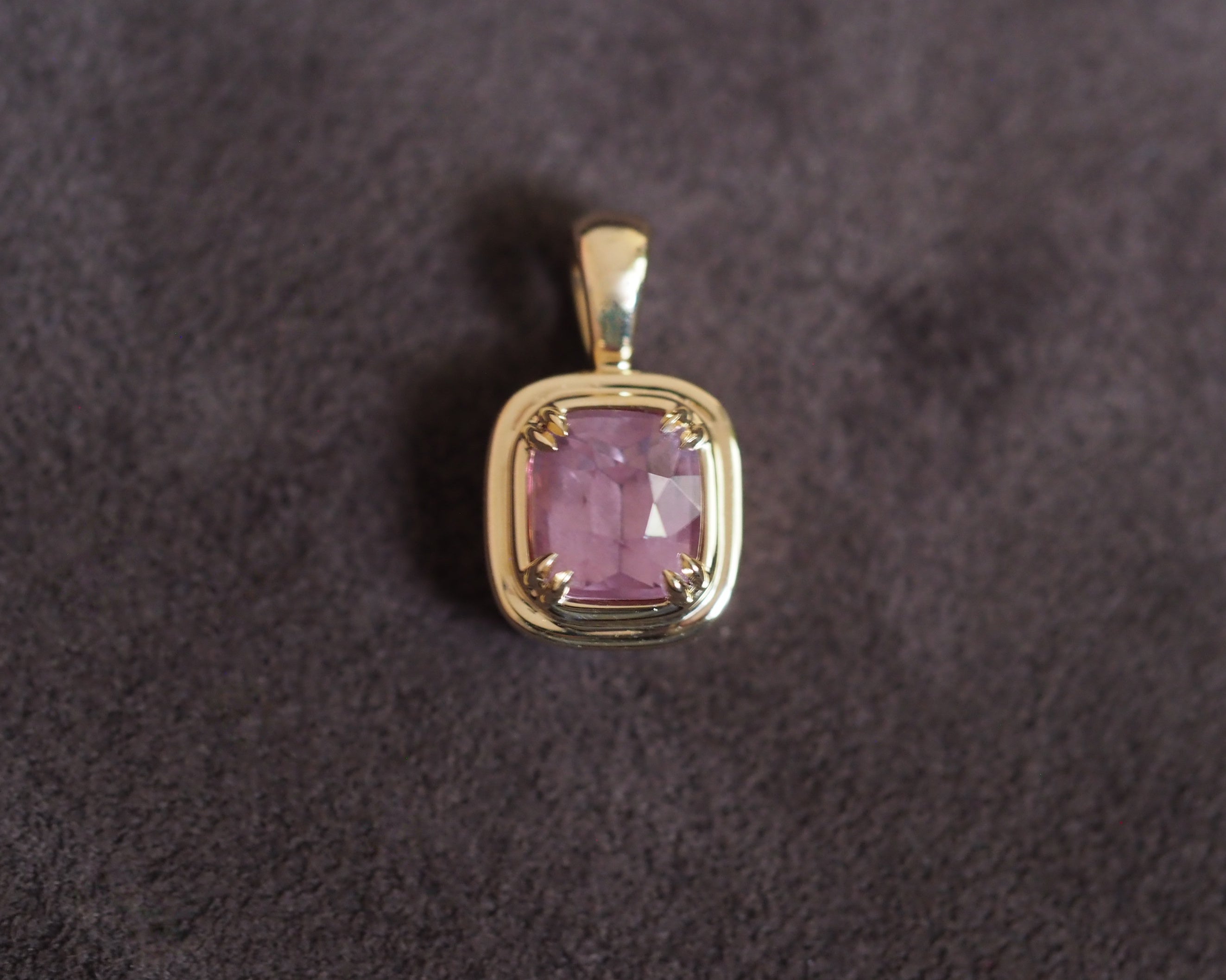 A gold pendant featuring a cushion-cut milky pink garnet, set against a soft, brown textured background.