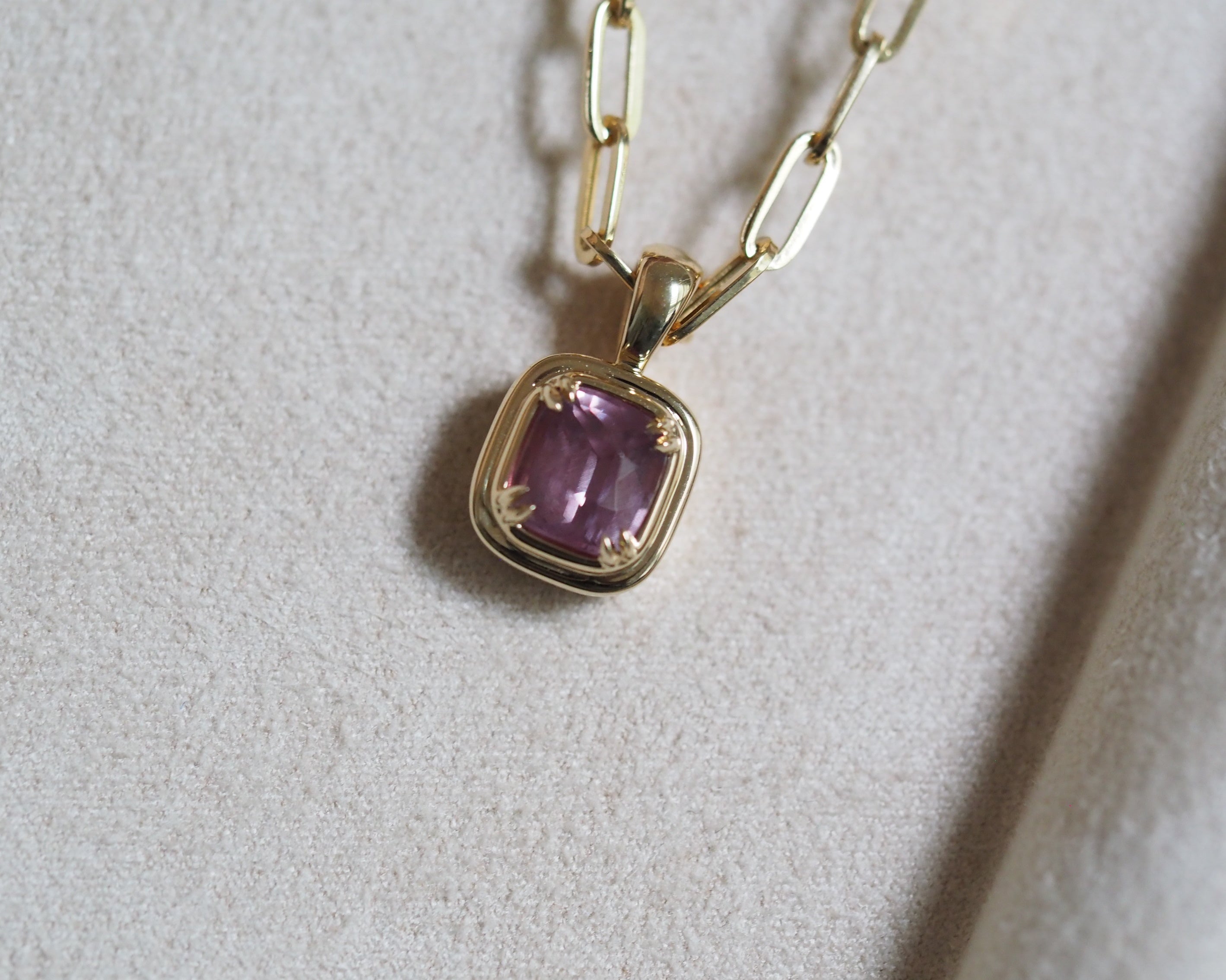 A gold pendant necklace featuring a Cushion-cut milky pink garnet in a gold stepped bezel setting, resting on a textured light gray surface.