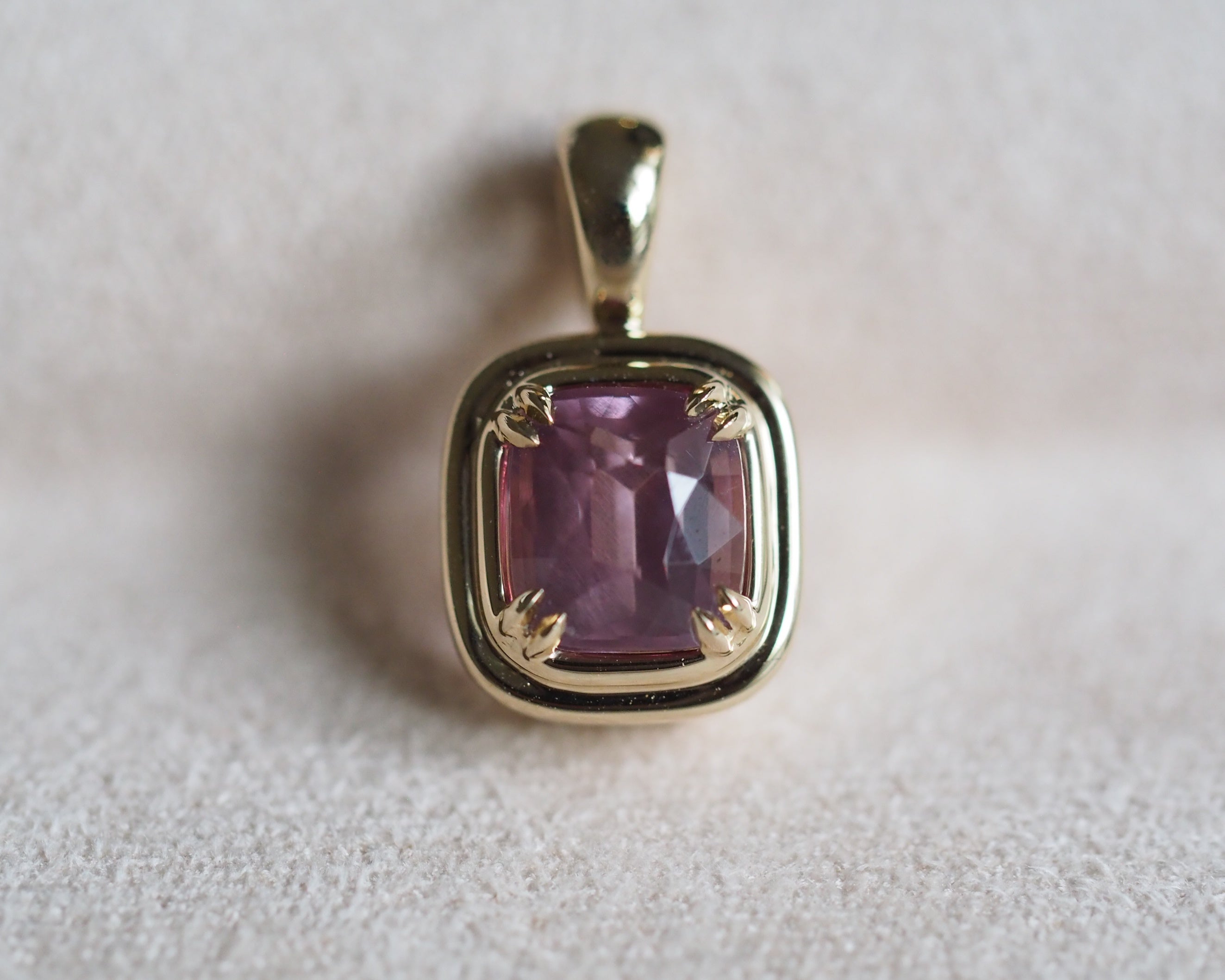 A gold pendant featuring a cushion-cut milky pink garnet, set against a soft, light beige textured background.