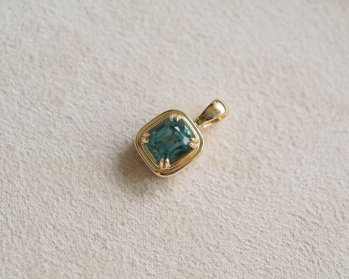 A gold pendant featuring a cushion-cut teal tourmaline in a prong setting, resting on a textured beige surface.