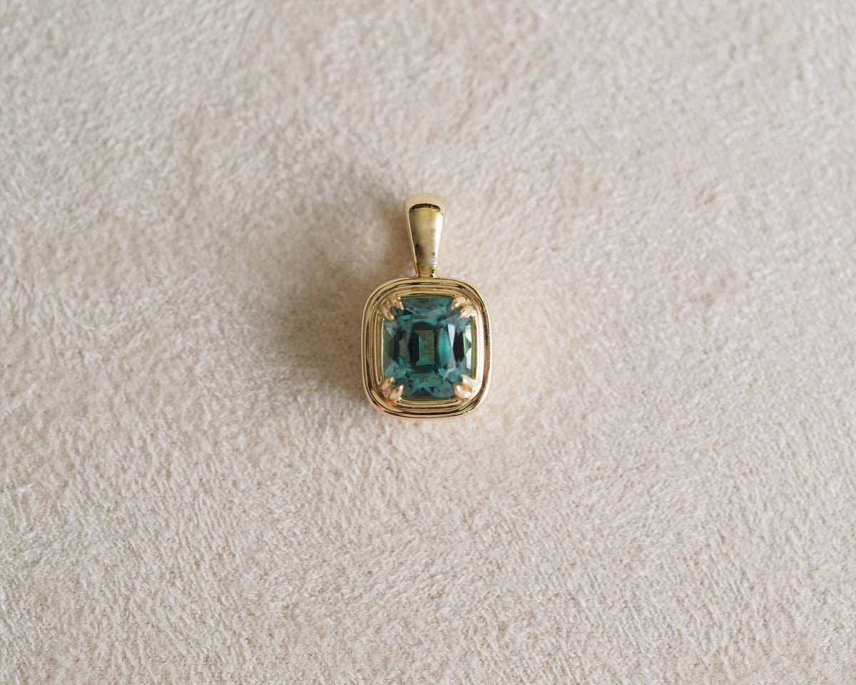 A gold pendant featuring a cushion-cut emerald-cut blue-green tourmaline, displayed on a textured beige surface.