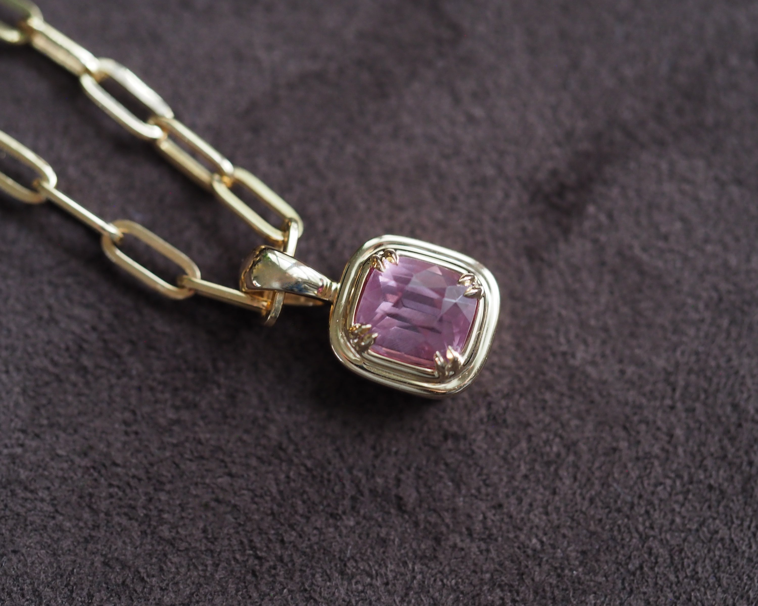 A gold chain necklace with a cushion bezel-set pink garnet pendant, displayed on a dark textured fabric background.