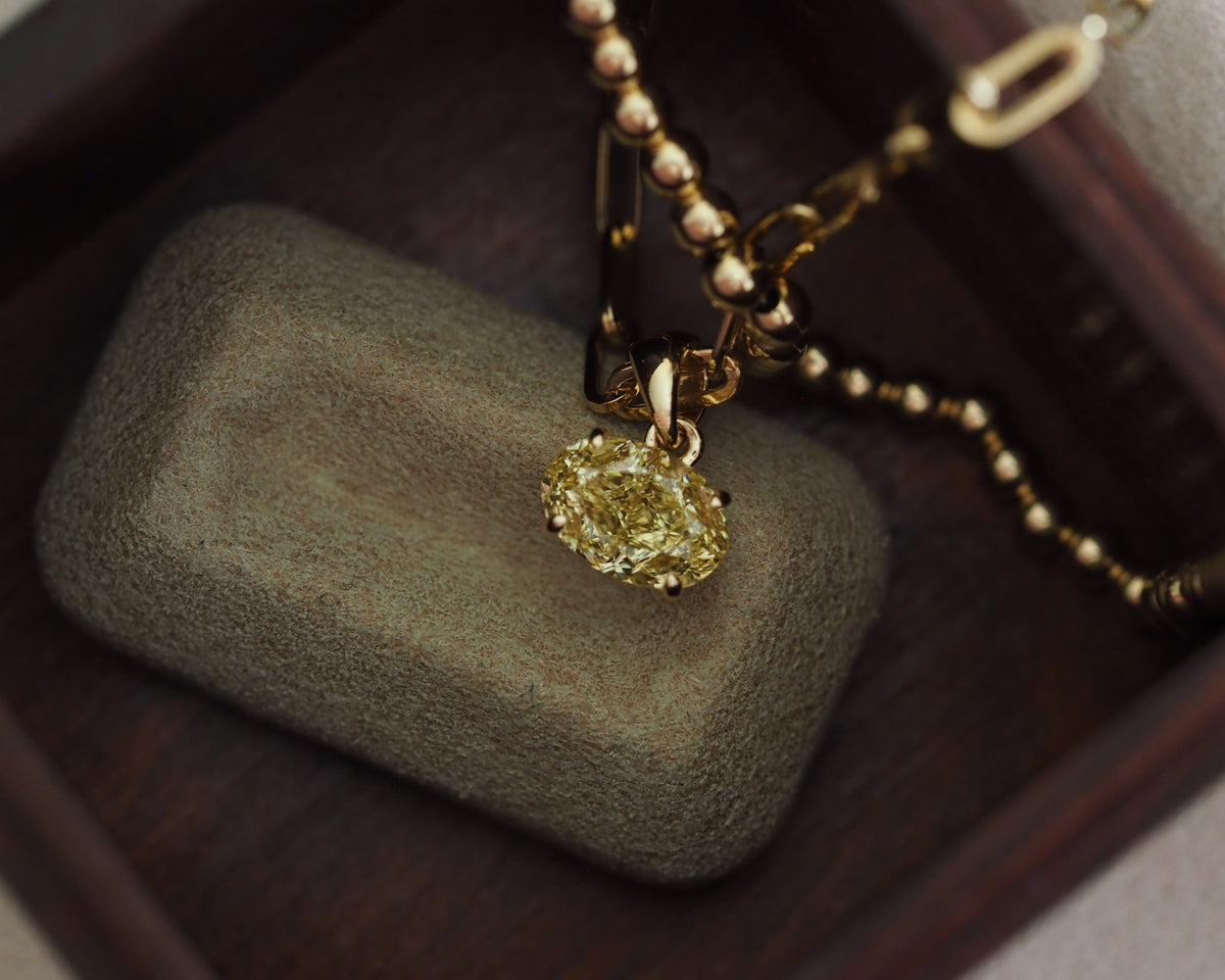 A yellow diamond pendant necklace in a gold setting with a gold beaded chain, resting on a textured gray stone inside a dark wooden jewelry box.