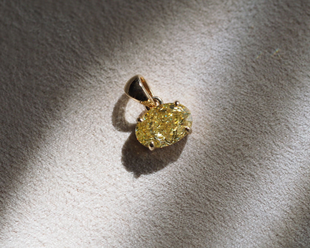 A yellow diamond pendant with a gold setting, resting on a textured beige surface under direct sunlight with a soft shadow.