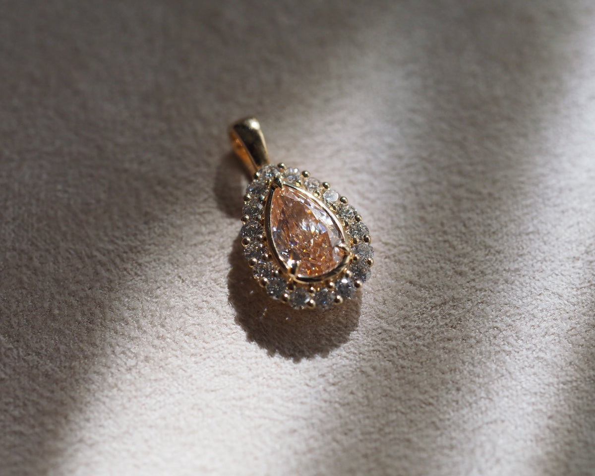 A pear-cut diamond pendant with a halo of small diamonds in a yellow gold setting, resting on a textured beige surface with dramatic light and shadow.