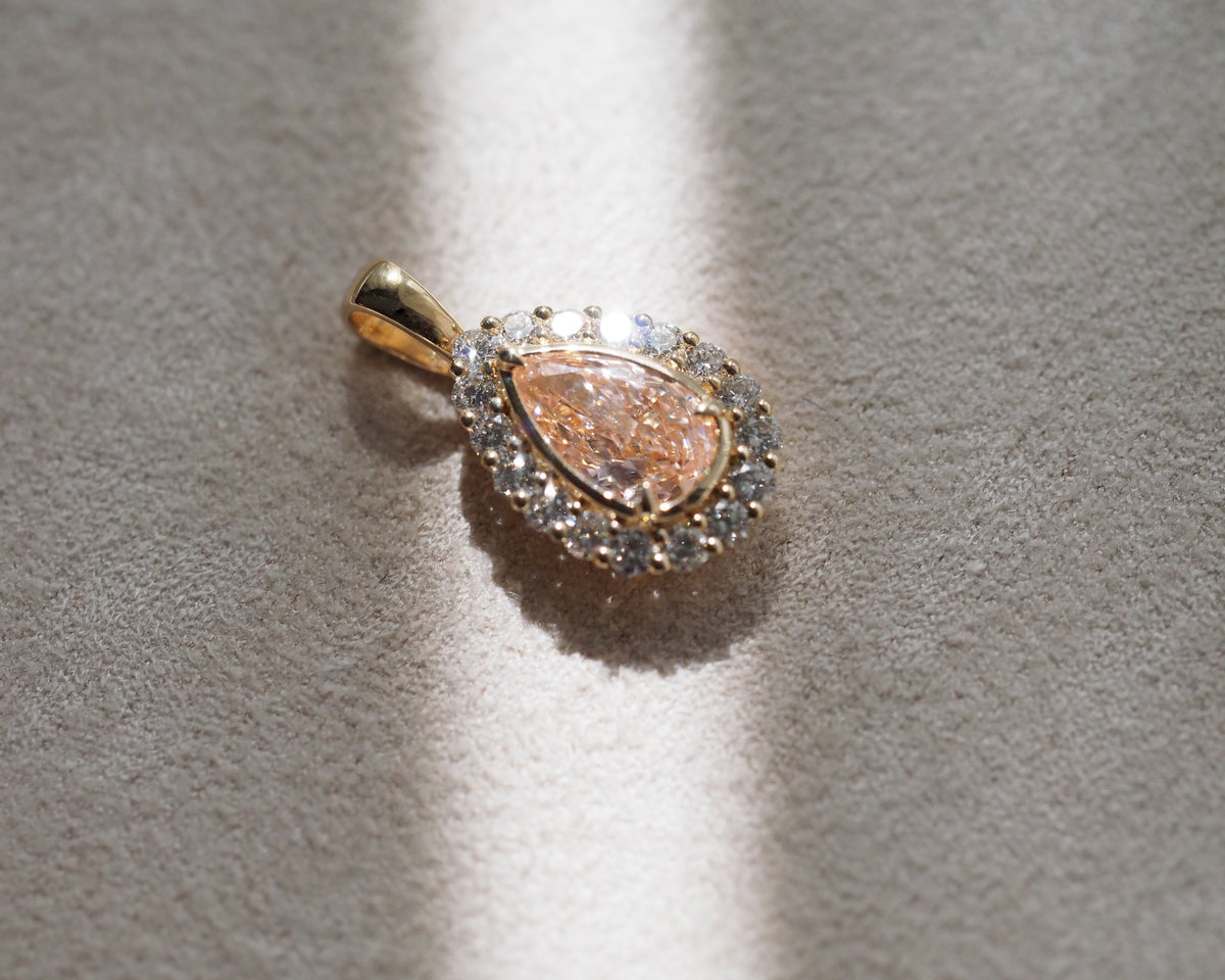 A gold pendant featuring a teardrop-shaped rose quartz center stone surrounded by a halo of small clear stones, displayed on a textured beige surface with direct sunlight and shadows.