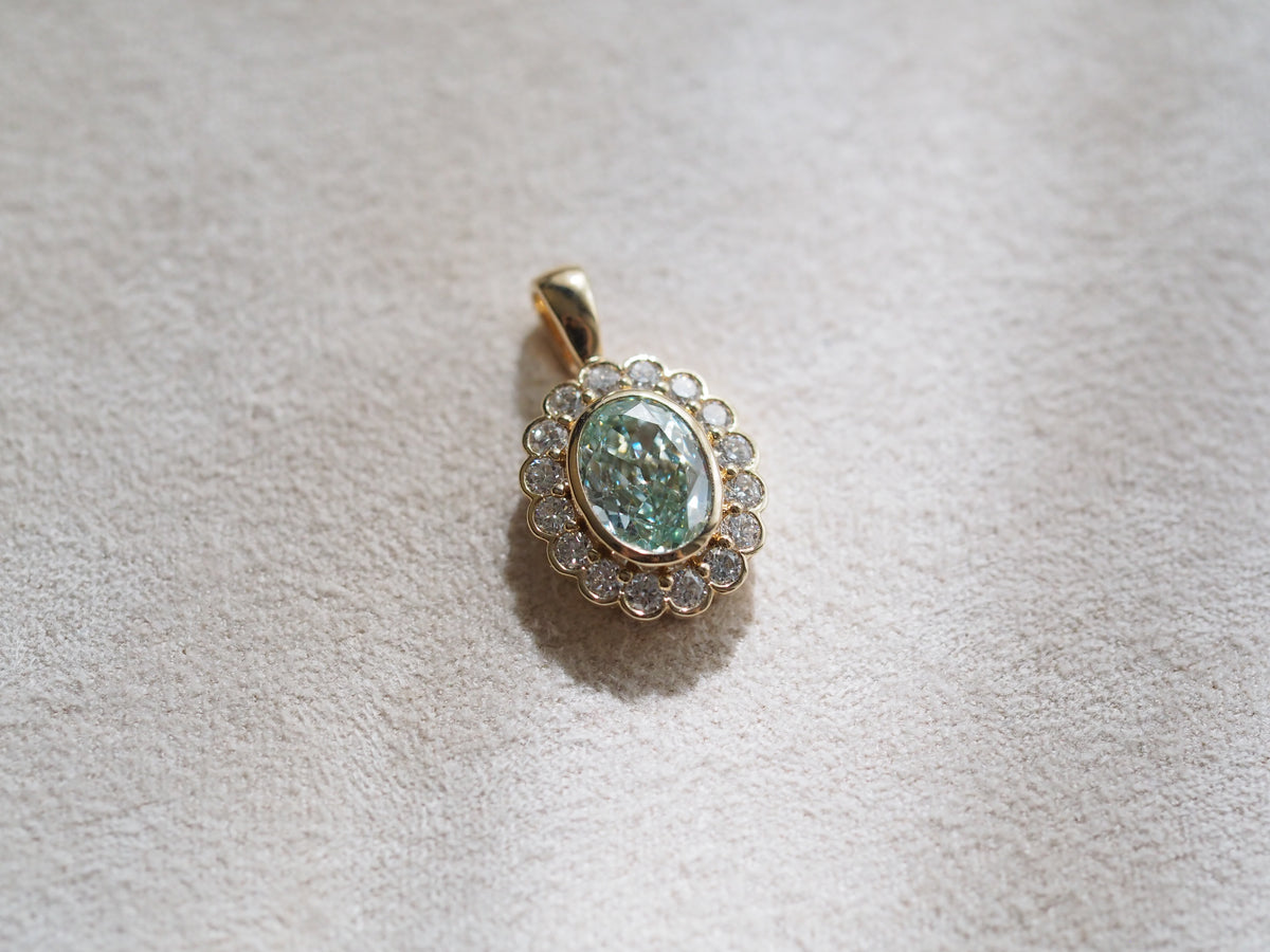 A gold pendant featuring an oval green stone surrounded by a halo of small clear stones, resting on a textured beige fabric surface.