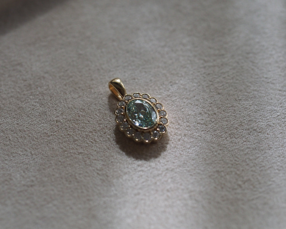 A gold pendant featuring an oval light blue-green gemstone surrounded by a halo of small clear stones, resting on a textured beige fabric surface.