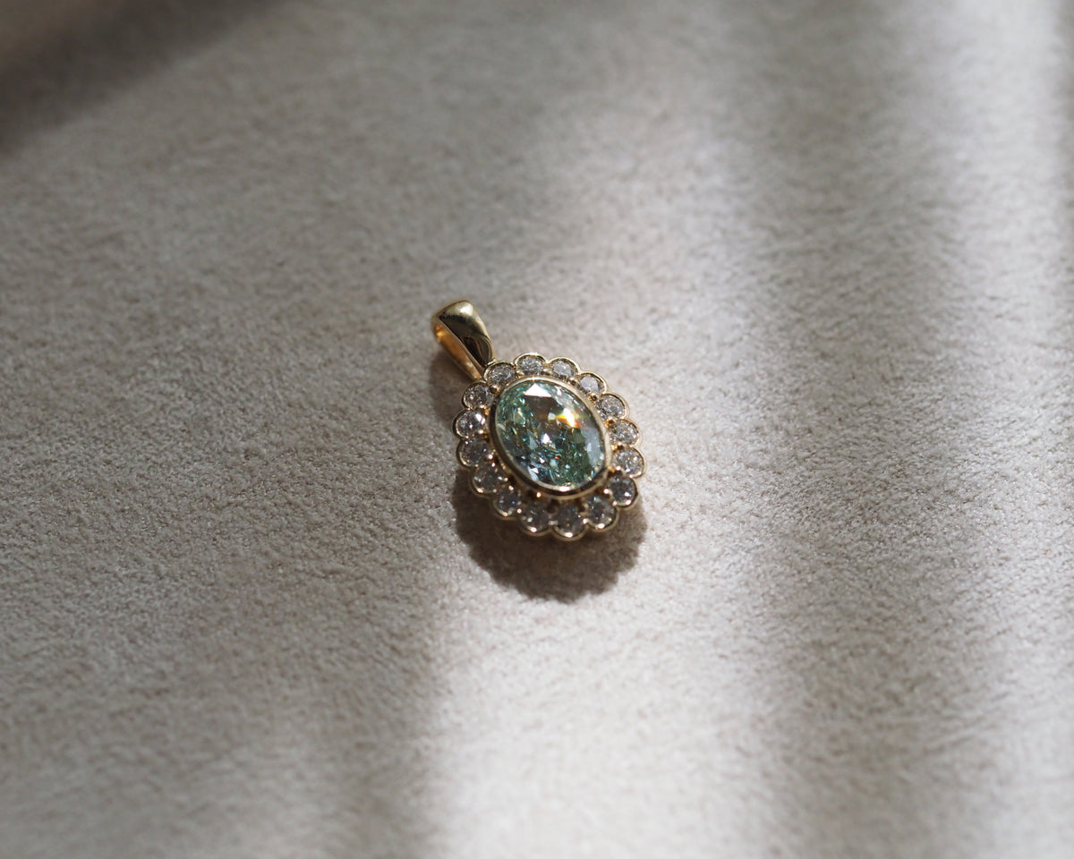 A gold pendant featuring a central oval light blue-green gemstone surrounded by a halo of small clear stones, resting on a textured beige fabric surface with soft shadows.