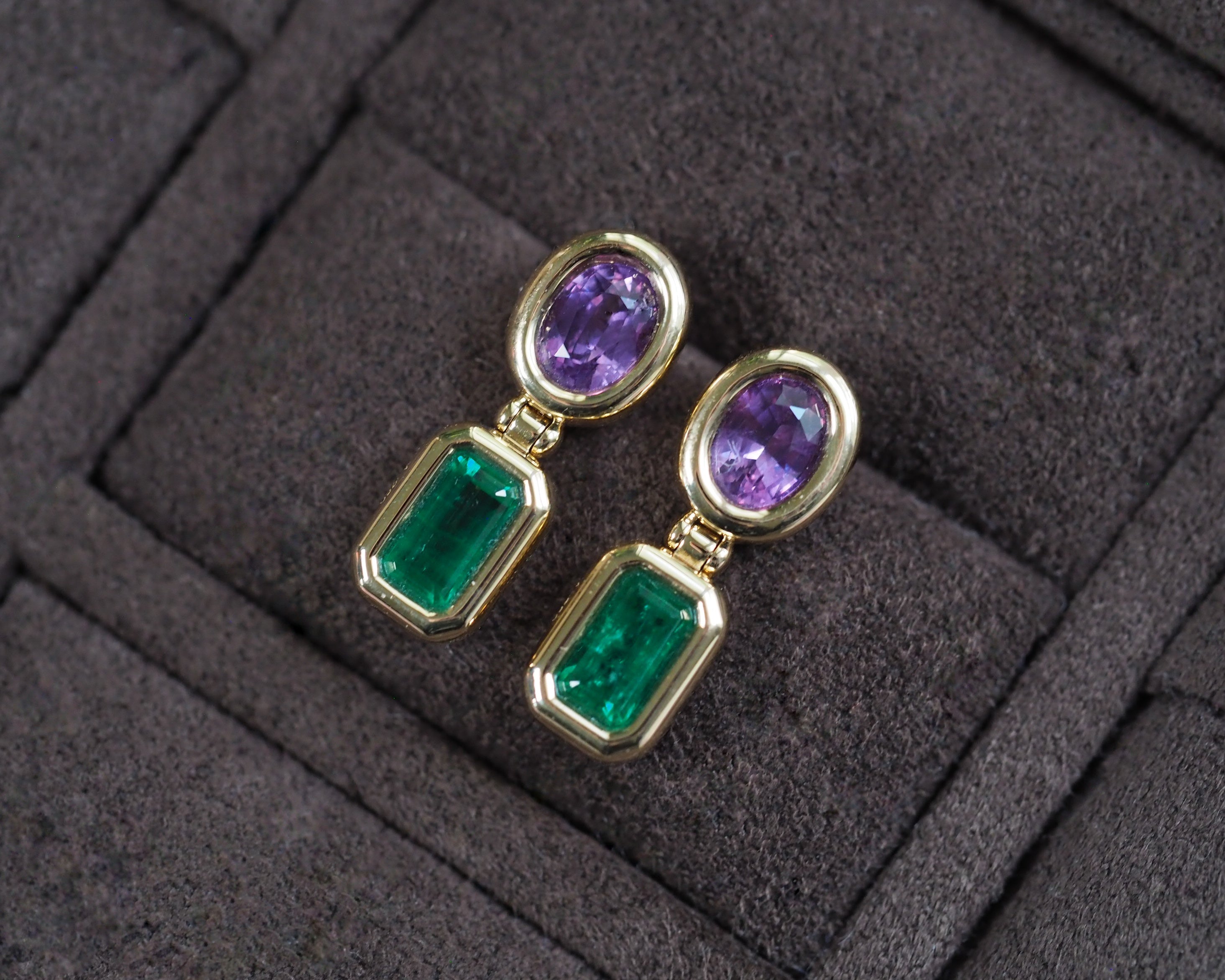 A pair of gold drop earrings with oval purple gemstones on top and rectangular emerald-cut green gemstones below, displayed on a dark textured velvet surface.