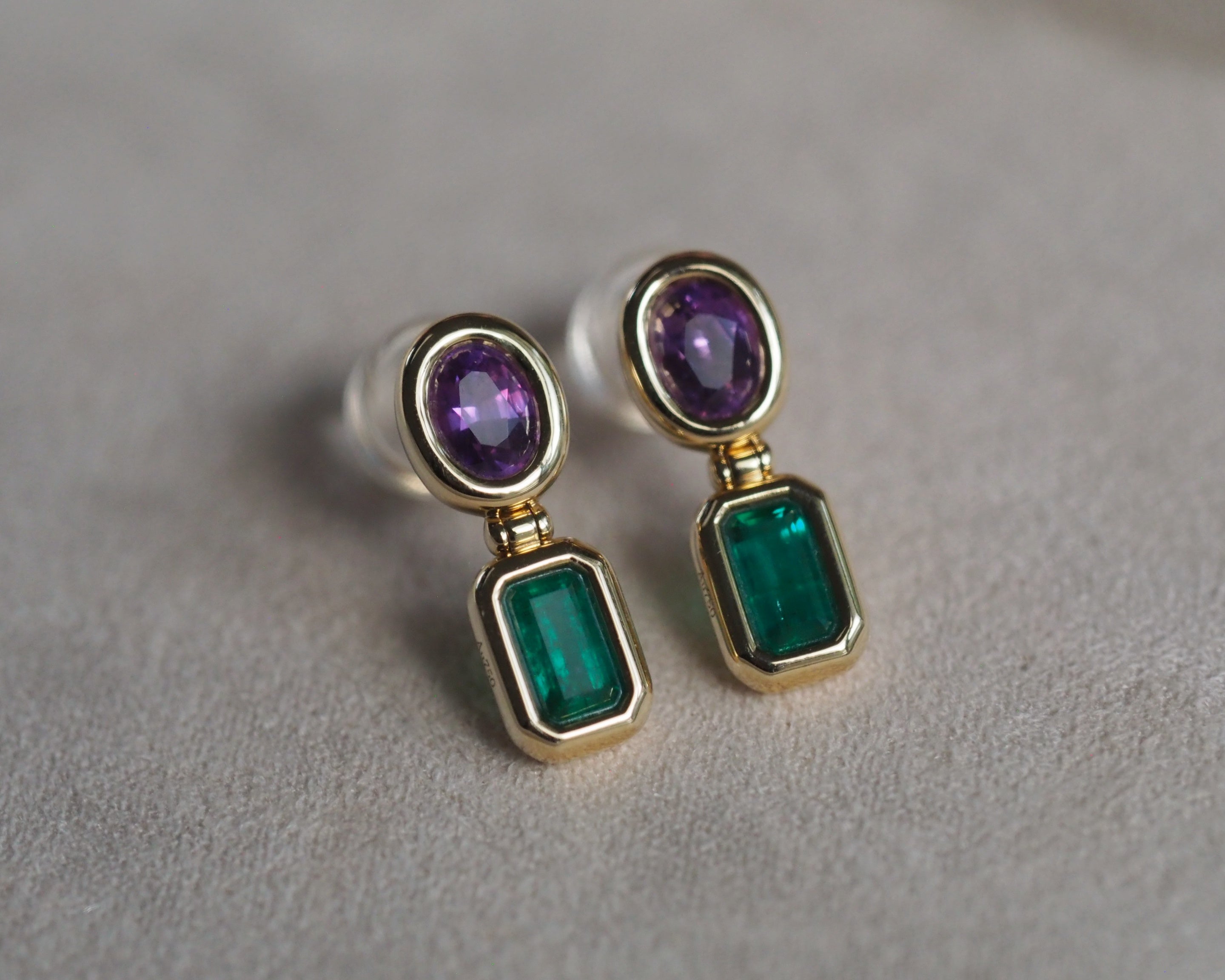 A pair of gold drop earrings featuring oval purple gemstones at the top and rectangular green emerald-cut gemstones below, displayed on a textured light gray surface.